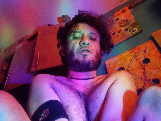 DavydWolfy69, zonder shirt, met krullend bruin haar, baard en een zwarte armband, zittend met de knieën opgetrokken, kijkend naar de camera, met een zwarte choker om, onder paars en groen licht..
