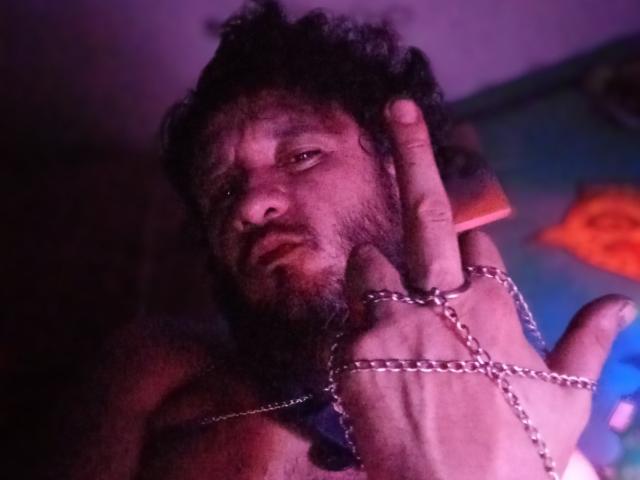 DavydWolfy69, com uma barba por fazer, cabelo escuro e desalinhado, e uma corrente no pulso, com uma expressão intensa, com o dedo levantado em direção à orelha, em um ambiente escuro, iluminado em tons de roxo..