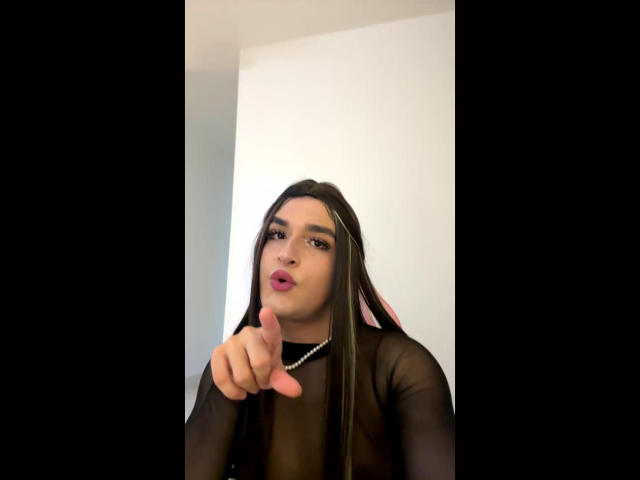 DaniBenson with long black hair, pouty pink lips, and a black sheer top, pointing a finger with a pink nail, wearing a pearl necklace.
