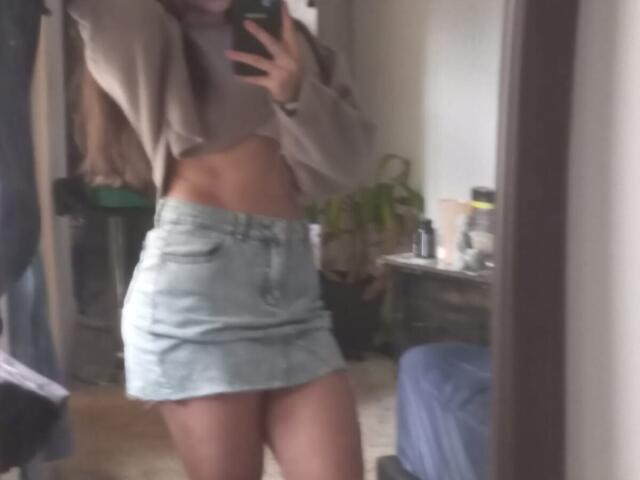 Ein Spiegelbild von SteffanyTaylor, die mit erhobenen Armen posiert, wobei ihr schlanker Bauch zu sehen ist. Sie trägt ein beiges Crop-Top und einen hellblauen Jeansrock und hält ein Handy..