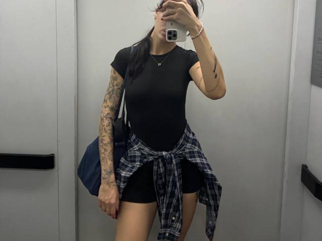 Tsukerberg with tattoos on her right arm, black shirt, black shorts, plaid shirt tied around her ass, holding a phone in front of her face, standing in a mirror.