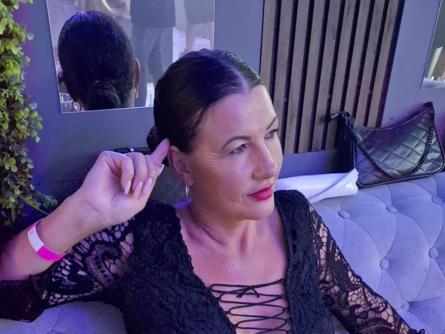FrancaiseAnnaDom with dark hair in a bun, wearing a black lace dress, red lipstick, adjusting her ear, sitting on a grey couch, wrist with pink bracelet.