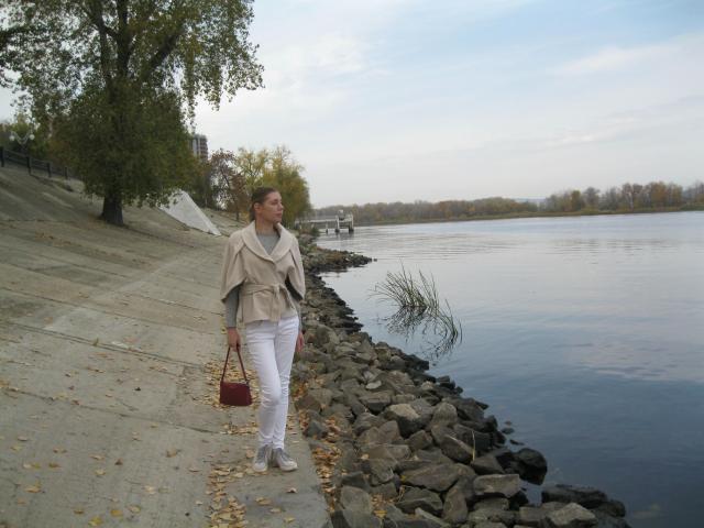 LoveFM-hot with beige shawl, white pants, and red purse walks by rocky water edge, holding grass, wearing white shoes, under tree with autumn leaves.