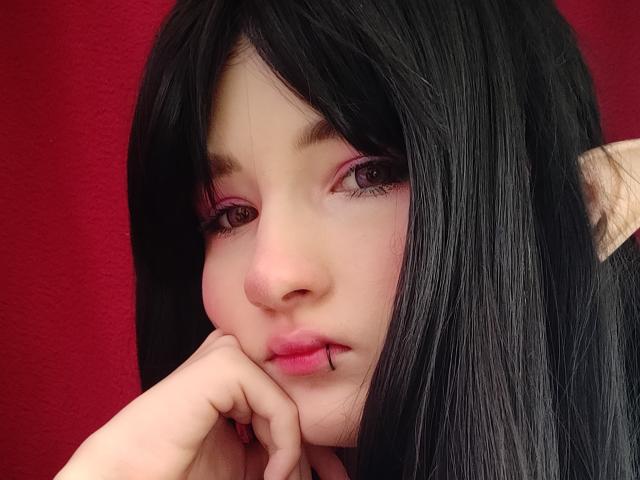 Selene Worrall tem cabelo preto, pálpebras rosadas, pele clara e um piercing no lábio com uma barra fina. Ela está tocando o queixo com a mão, parecendo um pouco triste..