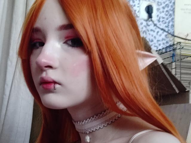 Selene Worrall, com cabelo laranja, pele clara, maquiagem rosa nos olhos, orelhas pontudas de elfo, um colar e uma pequena perfuração no nariz, olhando para a câmera com um leve semblante de descontentamento..