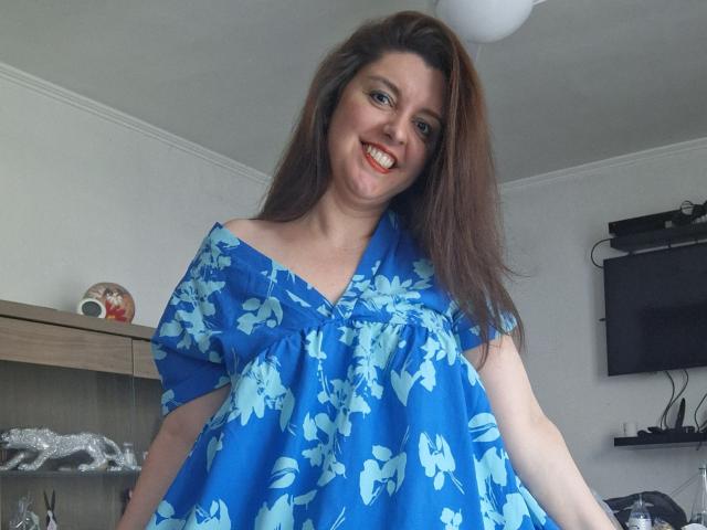 LaFrancaiseJessy with long brown hair, smiling, wearing a blue floral dress, exposing her big ass, standing in a room with a TV on the right and a wooden shelf.