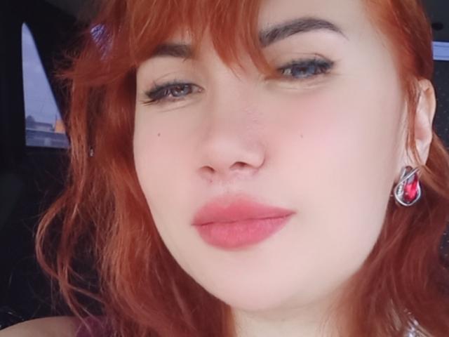 RoxolanaSexy has red hair, blue eyes, and pink lipstick. She's wearing pink earrings with red jewels. Her nose is slightly upturned, and she's giving a pouty look.