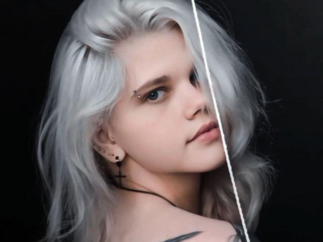JoannLowe with pale skin and platinum blonde hair, piercing above right eyebrow, black cross earring, looking to the side, white line partially covering face, bare shoulders with tattoo.
