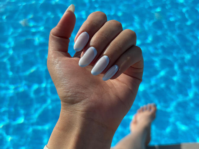 Pingli's hand with white, almond-shaped nails, showing off her fingers, in front of a blue, wavy pool. Her tan skin and a foot are visible in the background.