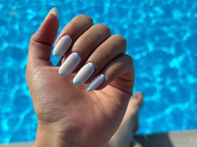 Pingli's hand with long, white, almond-shaped nails, holding up by a bright blue swimming pool. Her skin is tan, and her fingers are slightly curled. Her toes are visible.