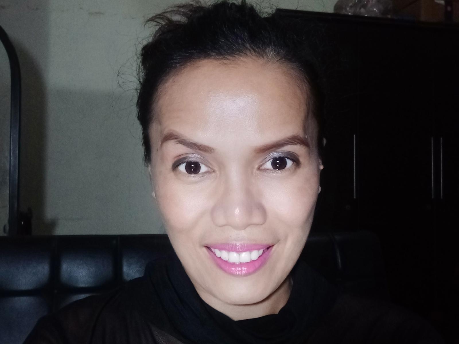 NikkiePhil has brown eyes, black hair in a messy updo, pink lipstick, and is smiling with white teeth. She's wearing a black sheer top.