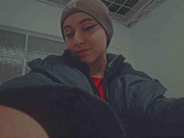 MissTat with a beanie, dark jacket, and red shirt, sporting winged eyeliner, a nose ring, and a slight smile, in a low-light selfie. Her ass is partially visible.