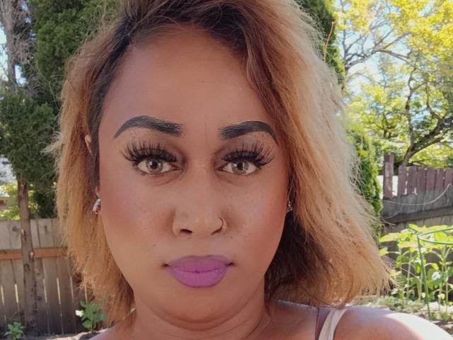 Aumerella's face with tan skin, purple lipstick, thick eyebrows, long eyelashes, nose ring, and shoulder-length blonde hair, wearing white top, standing outside in sunny garden.