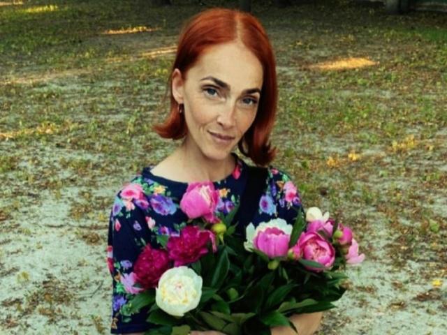 BreeTanya with red hair, holding pink and white peonies in a dark floral dress, smiling at the camera, standing on a grassy, leaf-strewn ground.