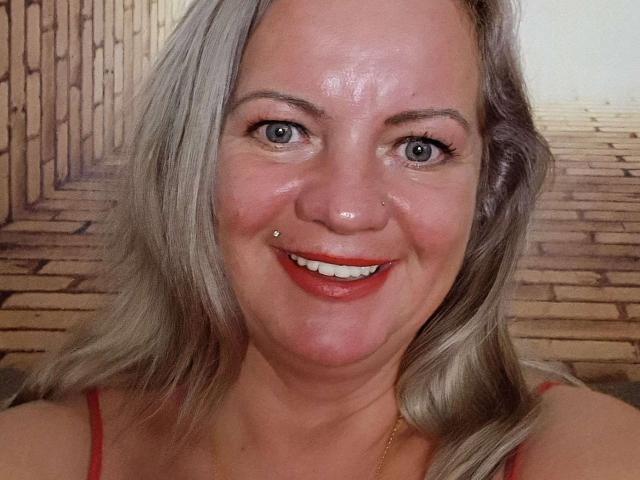 MolligSpielle has a smiling face with a nose piercing, red lipstick, blue eyes, and blonde hair, wearing a red top, with a shiny, slightly sweaty forehead.