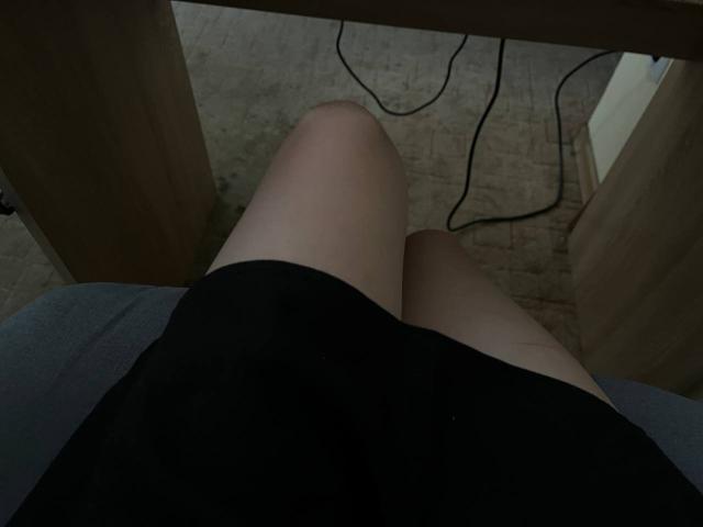 Valissia's onbedekte dijen onder een zwarte rok, benen lichtjes gespreid, met een houten tafel erboven en een zwarte kabel op een betegelde vloer. Valissia's achterkant is zichtbaar..