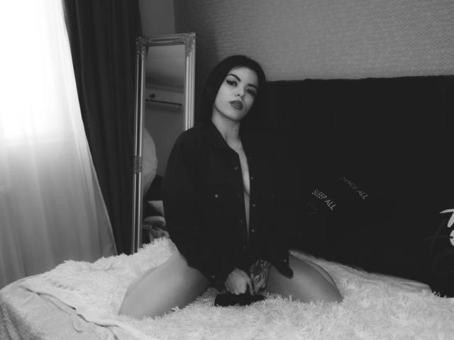 RinaQueenie with dark hair, wearing an open black shirt, kneeling on a bed with white fur, exposing her tits and holding a dildo, in a black and white photo.