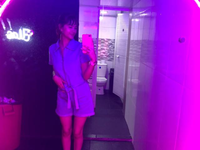 Melody Mansson de pie en un baño, vestida con un vestido azul, sosteniendo un teléfono, con luces de neón rosas y moradas que se reflejan en un espejo, mostrando su trasero y sus pequeños senos..