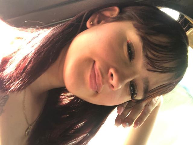 Melody Mansson, com cabelo castanho escuro, franja e um leve sorriso, com a cabeça apoiada na mão, mostrando uma tatuagem no ombro direito, com a luz do sol no rosto, luz suave..