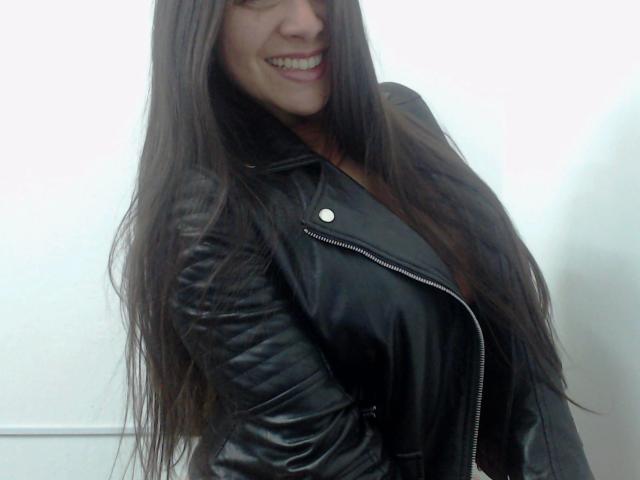 Gemma Lopez, attraente, con lunghi capelli castani, sorridente, indossa una giacca di pelle nera, mostra un piccolo bottone metallico e i suoi seni sono leggermente visibili sotto la giacca..