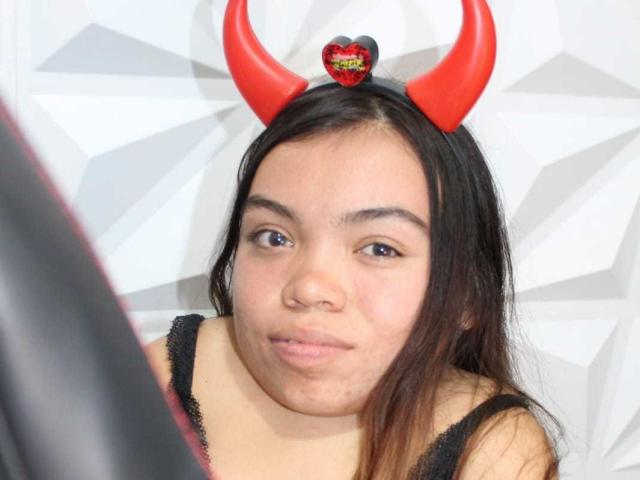 MichelleBrooks has red devil horns on her head, brown eyes, and is wearing a black lace top. Her expression is neutral with slightly pursed lips.