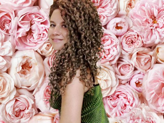 SelenaXCute with curly brown hair, green striped dress, smiling at camera, pink rose-filled background, cute and attractive, upper body, slight side view, showing tits and pretty face.