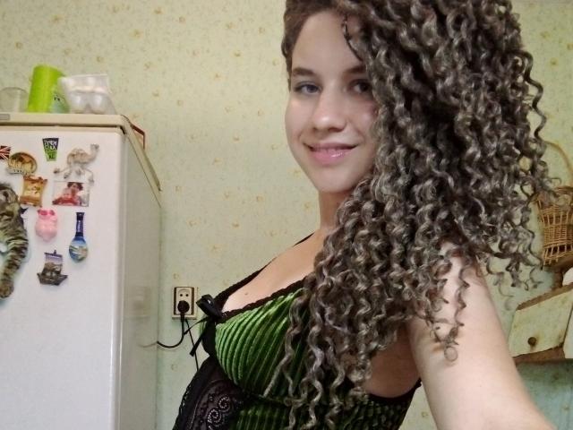 SelenaXCute with long curly brown hair, green and black striped lingerie, smiling, showing her tits, standing next to a white fridge in a kitchen.
