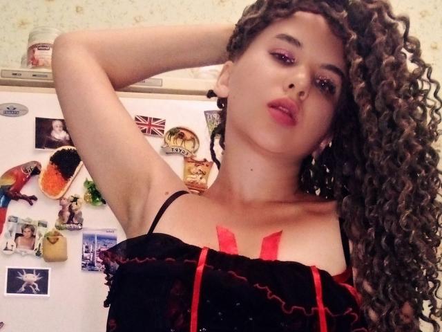 SelenaXCute with curly brown hair, red lipstick, and black/red lingerie, arms raised, showing off her tits with red straps. She's looking at the camera with a seductive expression.