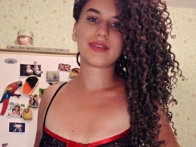 SelenaXCute with long curly brown hair covering one eye, wearing a black and red bra, smiling at the camera, standing in front of a white fridge.