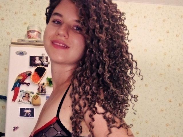 SelenaXCute with curly brown hair, smiling, wearing a black mesh bra with red trim, showing her tits, standing in a kitchen with a fridge and wall decor.