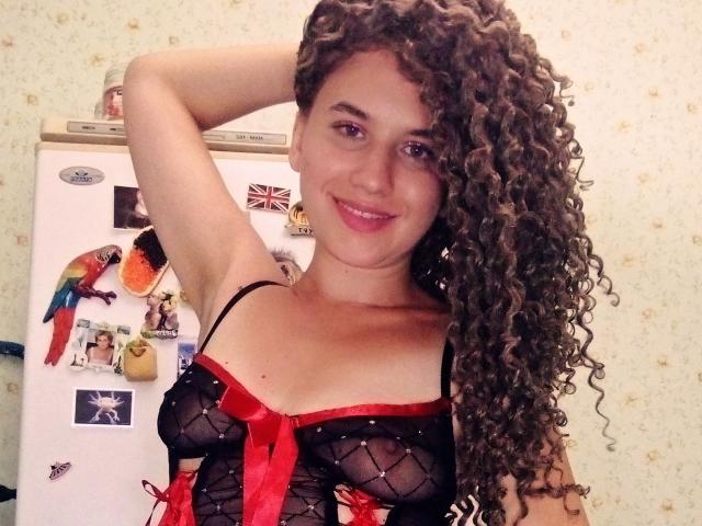 SelenaXCute with curly brown hair, smiling, wearing black sheer lingerie with red ribbon, exposing her small tits, has arm raised, standing in front of a fridge with a parrot.