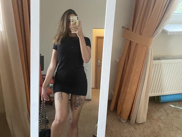 Kametia with a black dress, showing off her thighs with tattoos, taking a mirror selfie. Her phone has a floral pattern. She has brown hair, and her ass is cute.