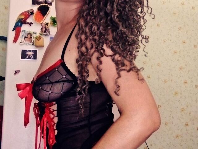 SelenaXCute with curly brown hair, wearing a black sheer lingerie top with red ribbon accents, showing her tits, standing sideways in a room.