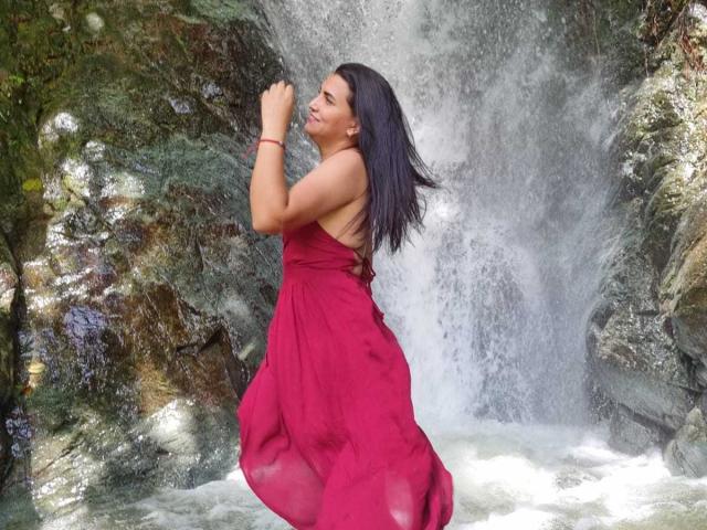 MarilynGarcia with long black hair, wearing a red dress, standing in front of a waterfall, eyes closed, smiling, holding her arm up, water splashing around her.
