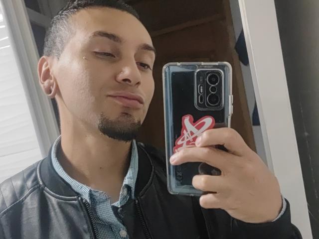 JohnDupontt with short black hair, light brown skin, and a goatee, wearing a black leather jacket and blue check shirt, taking a selfie with a red heart case.