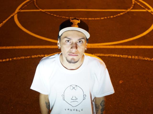 JohnDupontt with a white t-shirt, black cap, and tattoos, staring up with a weird expression, standing on a dark parking lot with yellow lines.