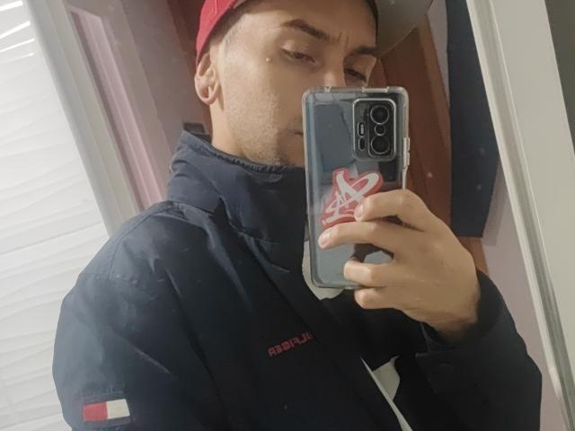 JohnDupontt taking a mirror selfie, wearing a red backward cap, black sport jacket, holding a phone with a heart sticker, slightly sweaty face, medium skin tone, short brown hair.
