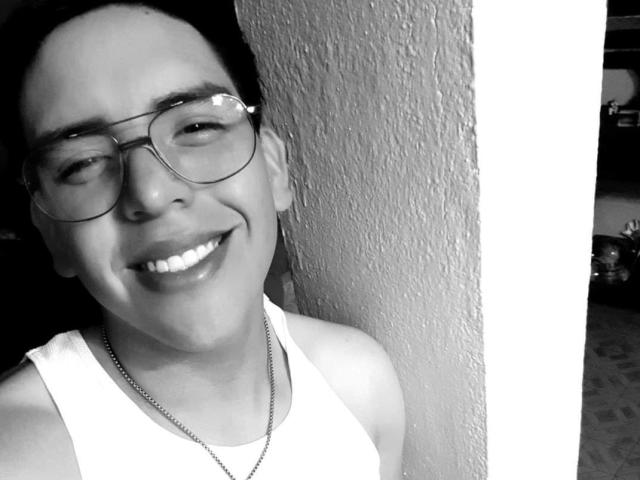 AnteRona with glasses, smiling, white tank top, necklace, standing by textured wall, black and white photo, looking slightly confused, medium build, short dark hair, visible teeth.