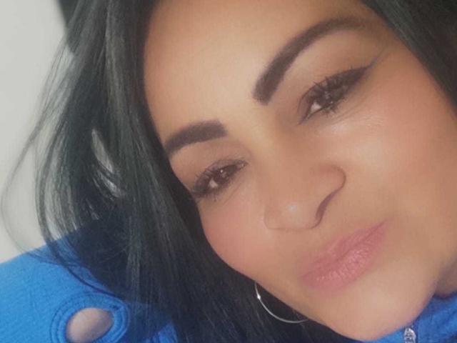 BeatriceTaylor has dark, straight hair, thick eyebrows, winged eyeliner, and pink lips. She's wearing a blue shirt and a silver hoop earring, with a subtle smile.
