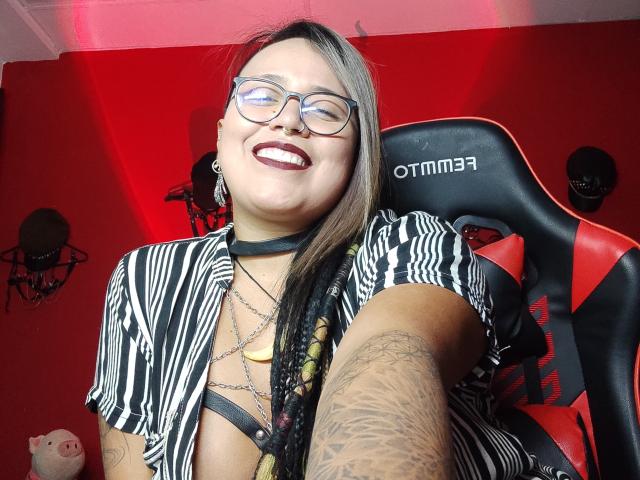 MistressLilith69 with glasses, dark lipstick, zebra-print shirt, black choker, tattoos, and braids, smiling, in a chair, showing her tits, red wall, and a black headset.