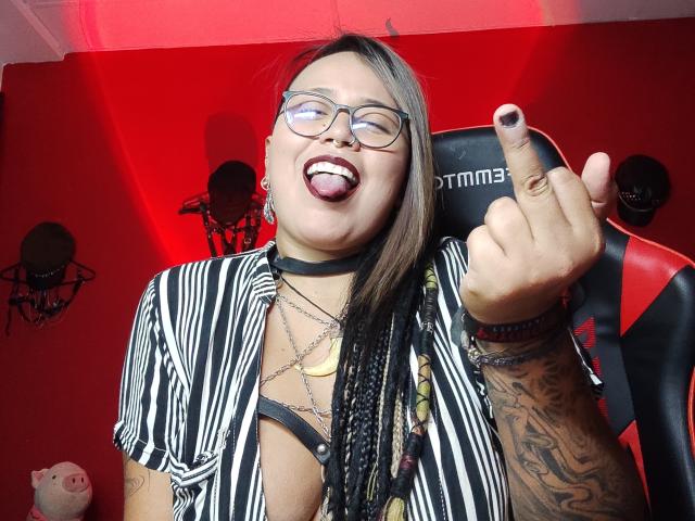MistressLilith69 with glasses, tongue out, and middle finger up, wearing a striped shirt, black choker, and tattoos, with braided hair and nipple piercings. Red room, gamer chair.