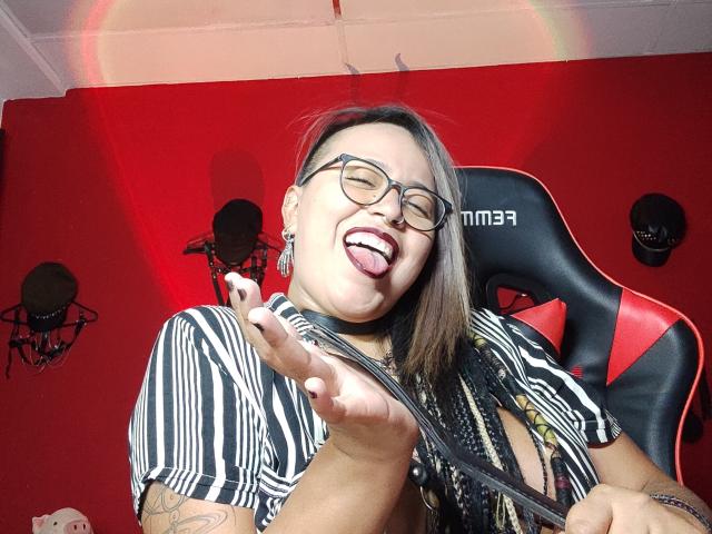 MistressLilith69 with tongue out, glasses on, striped shirt, black braids, tattoos, and a choker, holding a strap, smiling, in a red room with studio lights.