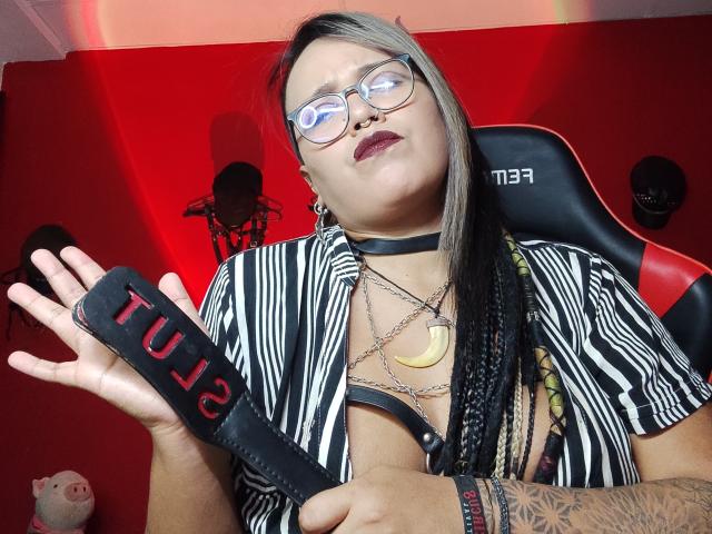 MistressLilith69 with glasses, striped shirt, black choker, and tattoos, holding a black leather paddle with "Tulip" in red, showing her tits and braided hair, red background.