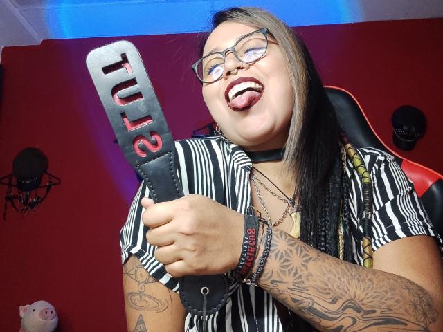 MistressLilith69, tongue out, holding a black and red flogga, wearing glasses, striped shirt, tattoos on arms, black choker, and multiple bracelets, in a chair.
