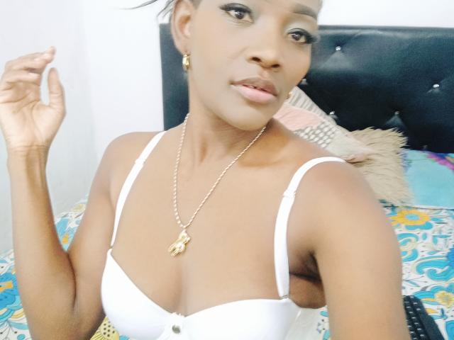 ToryMegans with dark skin, small tits in a white bra, gold necklace, earrings, and short hair, holding her hand up, looking at the camera, on a bed.