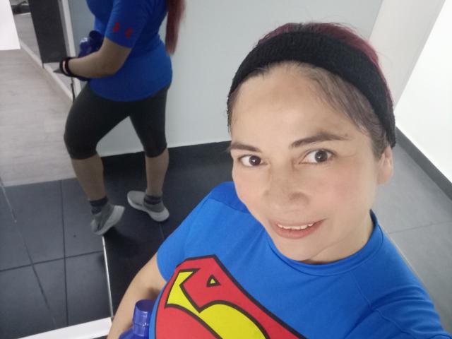 AlessiaRed with dark hair in a headband, blue Superman shirt, black leggings, and white sneakers, standing in front of a mirror, showing her reflection and smiling.