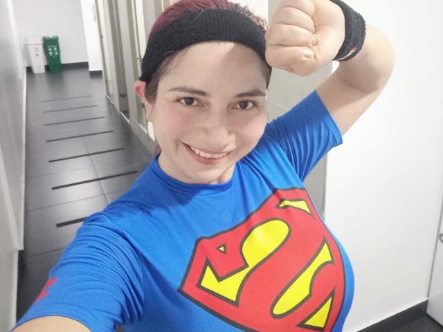 Alessia, cheveux roux avec des pointes rouges, bandeau noir, t-shirt Superman bleu, bras droit tendu, souriante, montrant un visage en forme, mignon, et de petites seins. Porte des bracelets noirs, fond blanc..