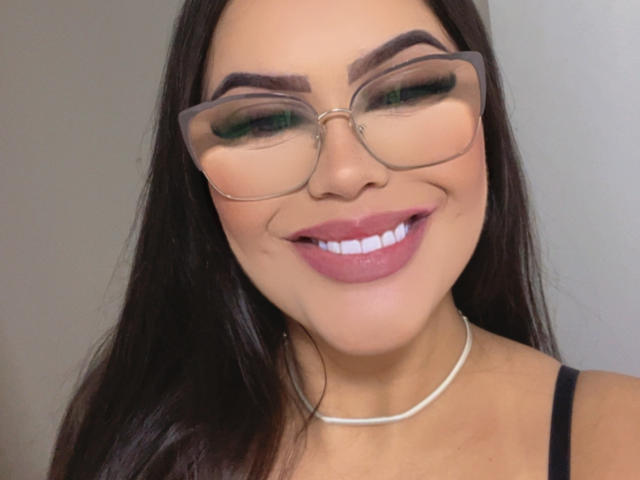 AnnneNaughtyBusty with glasses, dark eyebrows, green eyeshadow, pink lipstick, smiling, white necklace, black tank top, long dark brown hair, light skin.