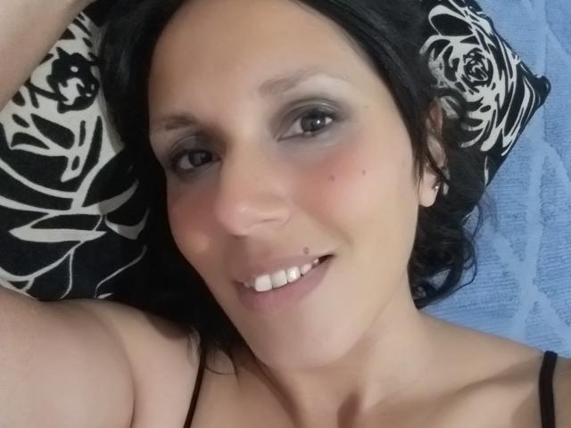  AlexandraSosa's face with black hair, grey eyeshadow, small nose piercing, smiling, showing white teeth, on a black and white rose pillow, wearing a black strap top.