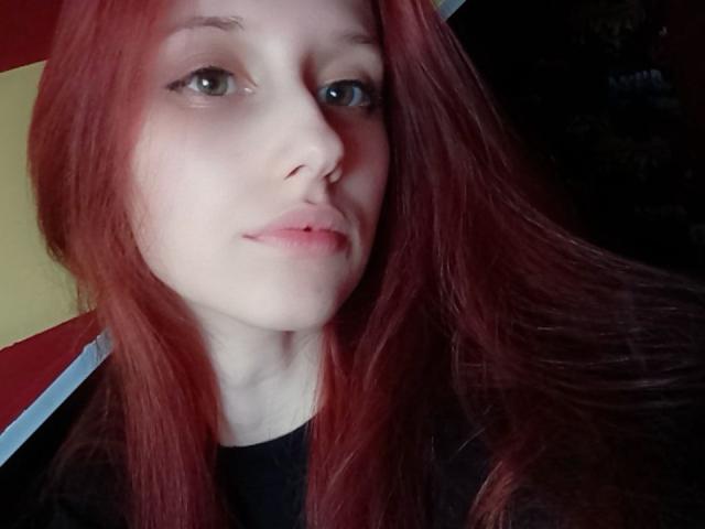 LunaXKiss has pale skin, red straight hair, green eyes, and a small nose. Her pink lips are slightly parted, wearing a black shirt, in a dimly lit setting.